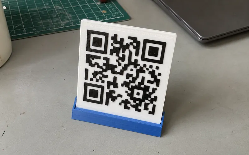 White raised QR code plaque in a blue printed stand on a maker desk.